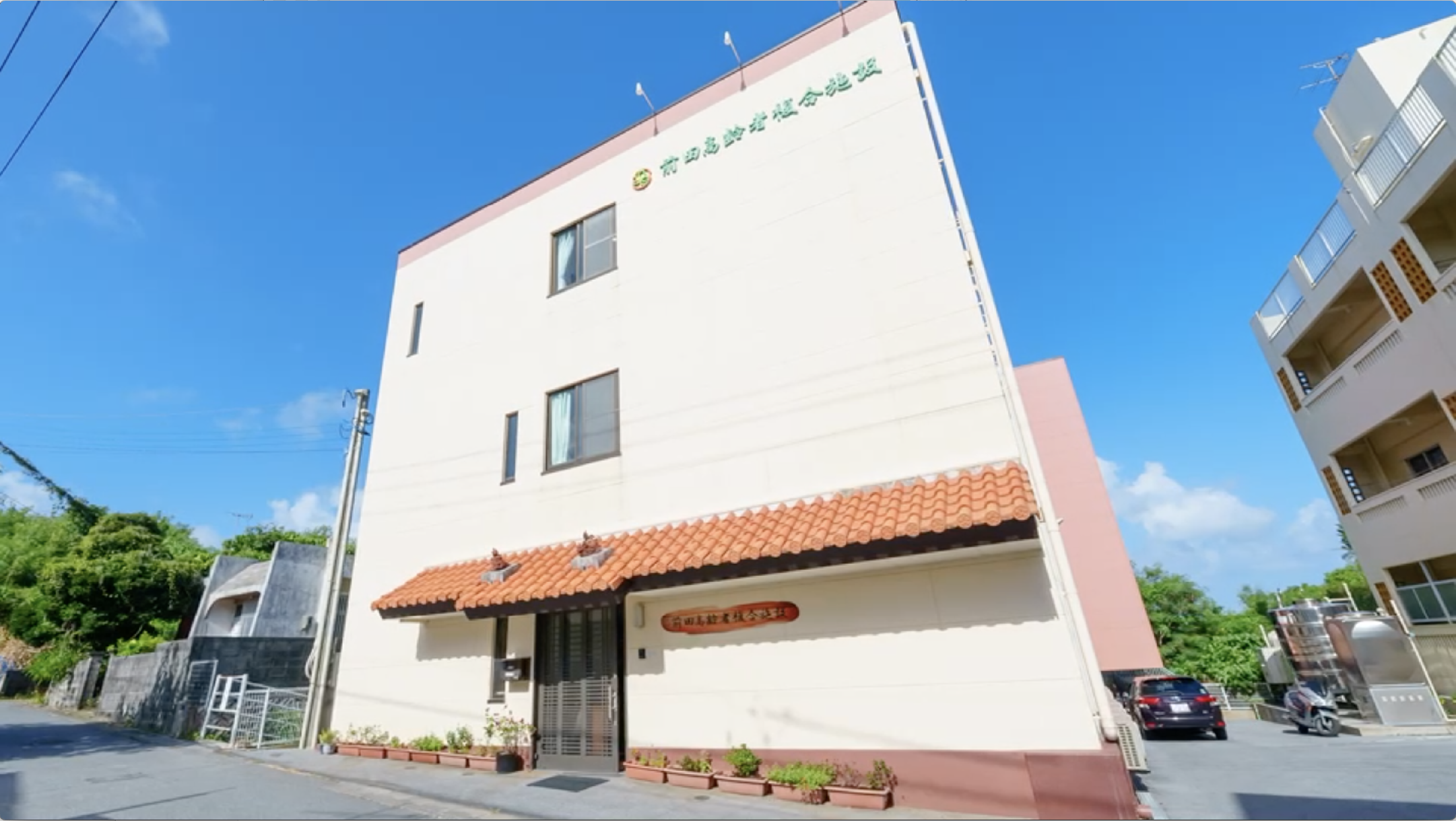 白と薄紅色の2階建て介護施設の外観。晴天下で周辺環境が明確に見える。